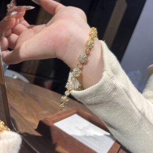 Könnte beinhalten: Ein goldfarbenes Armband mit floralem Design und klaren, funkelnden Edelsteinen. Das Armband besteht aus mehreren blütenförmigen Gliedern, die durch kleinere, kreisförmige Glieder verbunden sind. Es wird am Handgelenk getragen und zeigt seine filigranen Details.