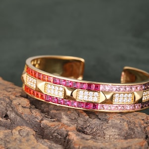 May include: Gold cuff bracelet with a geometric design. The bracelet features alternating rows of red, pink, and white gemstones. The gemstones are set in a gold band. The bracelet is displayed on a piece of wood.