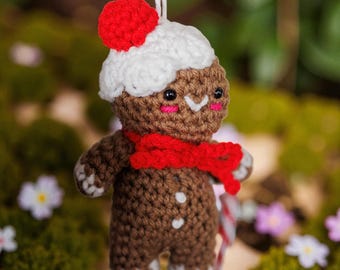 May include: A handmade crochet gingerbread man ornament. The brown gingerbread man has a white hat with a red pom-pom, a red scarf, and white accents. The ornament is suspended by a white string and has a candy cane. The background is blurred.