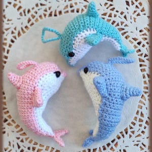 Puede incluir: Tres delfines hechos a mano en crochet, en tonos azul claro, rosa y azul, con vientres blancos y ojos negros. Los delfines están dispuestos en un patrón circular, mostrando su detallado tejido y diseño juguetón. Un artículo artesanal encantador.