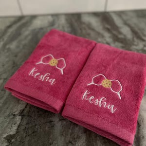 May include: Two pink towels with white embroidery. The towels feature a cookie-shaped bow and the name "Kesha" embroidered on each towel.