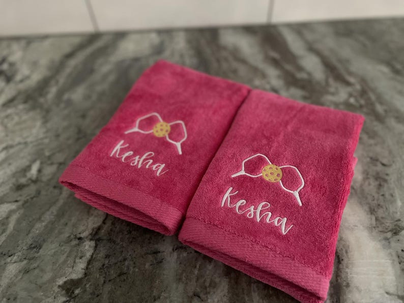 May include: Two pink towels with white embroidery. The towels feature a cookie-shaped bow and the name "Kesha" embroidered on each towel.
