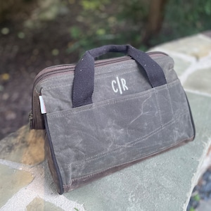 May include: A waxed canvas bag in shades of gray and brown, featuring a black handle and a front pocket. The bag has a zipper closure and a white embroidered monogram "C|R" on the front. The bag is designed for carrying items.