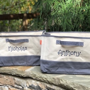 May include: Two canvas storage bins with gray bottoms and handles. The bins are beige with the names "Nicholas" and "Anthony" embroidered in black. Small orange tags are on the sides. The bins are sitting on a stone wall.