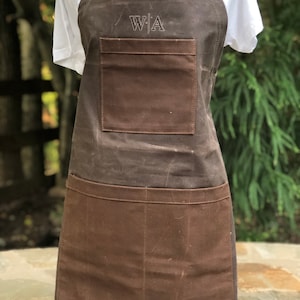 May include: A dark brown waxed canvas apron featuring a single chest pocket and a large double-compartment lower pocket. The chest area is embroidered with the letters W|A. The apron includes dark brown fabric neck and waist straps for securing.