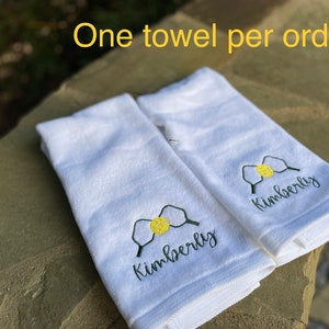 May include: Two white towels with the name "Kimberly" embroidered on them in black cursive lettering. Each towel features a green and yellow embroidered image of a pickleball paddle and ball. The text "One towel per order" is visible in the background.