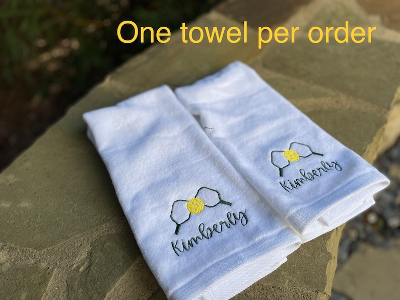 May include: Two white towels with the name "Kimberly" embroidered on them in black cursive lettering. Each towel features a green and yellow embroidered image of a pickleball paddle and ball. The text "One towel per order" is visible in the background.