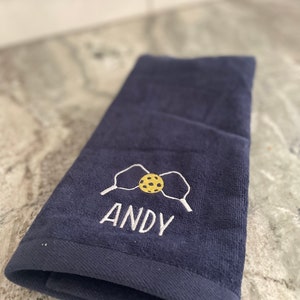 May include: A dark blue hand towel with white embroidery of two paddles and a yellow and brown ball. The name "ANDY" is embroidered below the paddles and ball.