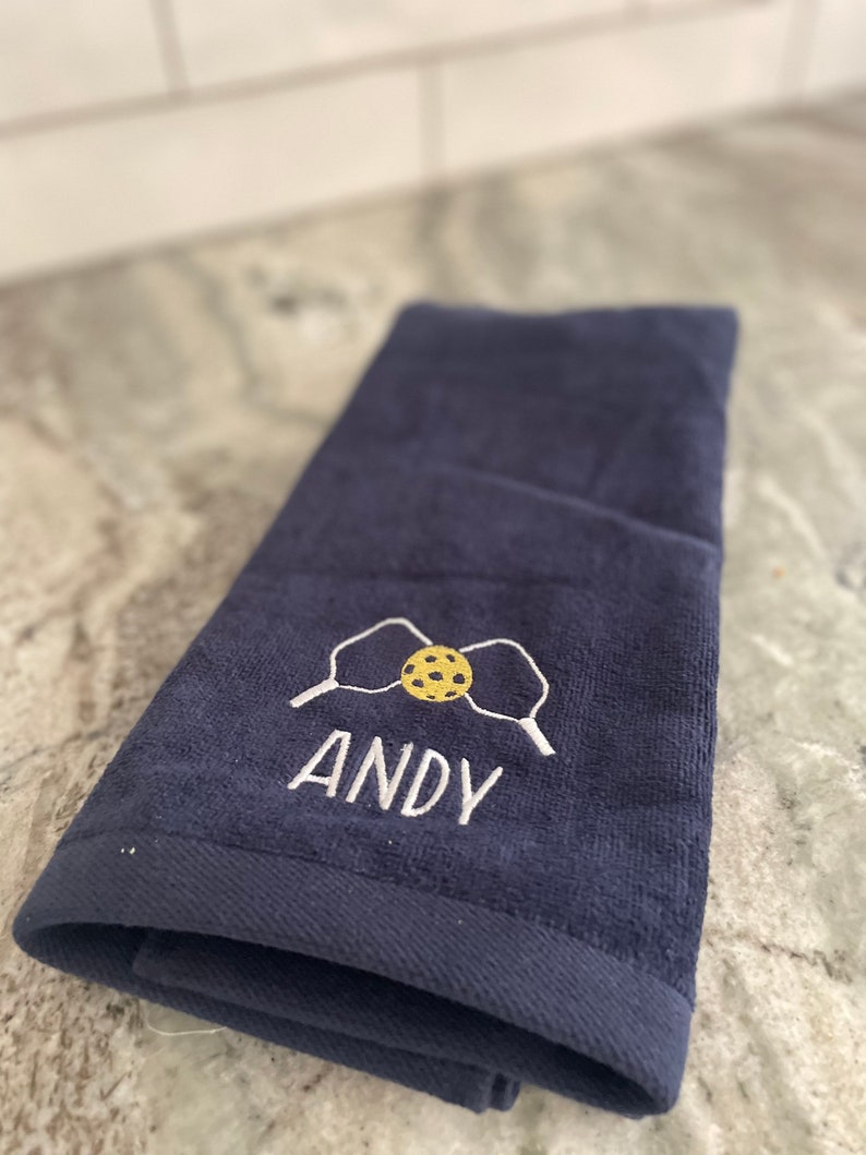 May include: A dark blue hand towel with white embroidery of two paddles and a yellow and brown ball. The name "ANDY" is embroidered below the paddles and ball.