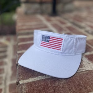 May include: A white sun visor featuring an embroidered rectangular patch of the United States flag on the front center. The visor has a curved brim and a structured headband design with visible stitching along the edges of the fabric.