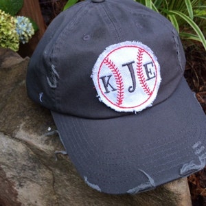 K&ouml;nnte beinhalten: Eine verwaschene, graue Baseballkappe mit einem Baseball-Patch mit den Initialen "KJE" in schwarzer Stickerei. Der Baseball-Patch hat rote N&auml;hte und einen wei&szlig;en Hintergrund. Die Kappe hat einen Used-Look.