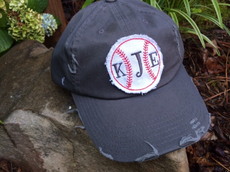 K&ouml;nnte beinhalten: Eine verwaschene, graue Baseballkappe mit einem Baseball-Patch mit den Initialen "KJE" in schwarzer Stickerei. Der Baseball-Patch hat rote N&auml;hte und einen wei&szlig;en Hintergrund. Die Kappe hat einen Used-Look.