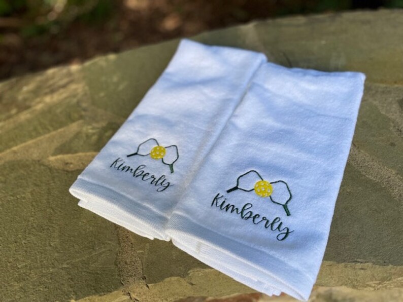 May include: Two white towels with embroidered yellow and green pickleball paddles and the name "Kimberly" in black cursive.