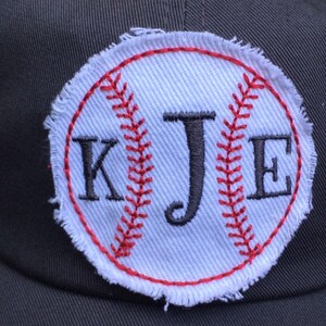 K&ouml;nnte beinhalten: Dunkelgraue Baseballkappe mit einem wei&szlig;en Baseball-Patch. Der Patch zeigt rote N&auml;hte und die Initialen "KJE" in Schwarz. Der Baseball-Patch hat einen ausgefransten, ausgefransten Rand.
