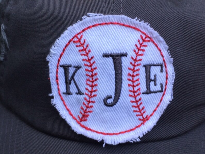 K&ouml;nnte beinhalten: Dunkelgraue Baseballkappe mit einem wei&szlig;en Baseball-Patch. Der Patch zeigt rote N&auml;hte und die Initialen "KJE" in Schwarz. Der Baseball-Patch hat einen ausgefransten, ausgefransten Rand.