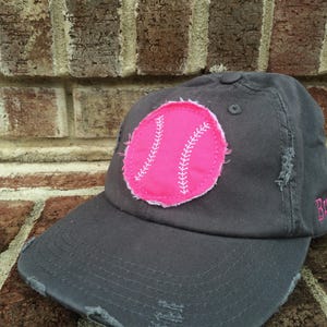 K&ouml;nnte beinhalten: Graue Baseballkappe mit abgenutztem Schirm und einem rosa Baseball-Patch. Der Patch hat wei&szlig;e Nahtdetails. Das Wort "Brave" ist in Rosa auf die Seite der Kappe gestickt.