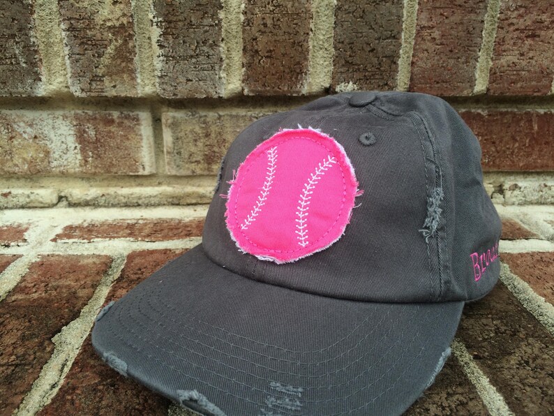 K&ouml;nnte beinhalten: Graue Baseballkappe mit abgenutztem Schirm und einem rosa Baseball-Patch. Der Patch hat wei&szlig;e Nahtdetails. Das Wort "Brave" ist in Rosa auf die Seite der Kappe gestickt.