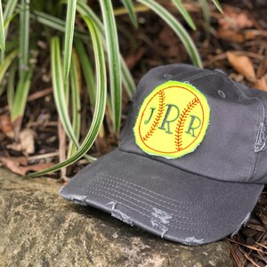 K&ouml;nnte beinhalten: Anthrazitgraue Baseballkappe mit abgenutztem Rand. Die Kappe hat einen gelben Aufn&auml;her mit Softball-Design und den gestickten Initialen "JPR" in Gr&uuml;n.