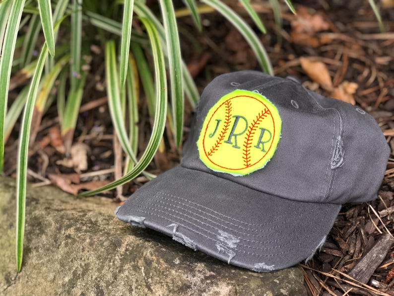 K&ouml;nnte beinhalten: Anthrazitgraue Baseballkappe mit abgenutztem Rand. Die Kappe hat einen gelben Aufn&auml;her mit Softball-Design und den gestickten Initialen "JPR" in Gr&uuml;n.