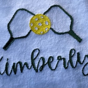 May include: A white towel with green embroidered pickleball paddles and a yellow ball in the center. The name Kimberly is embroidered below the paddles in green.