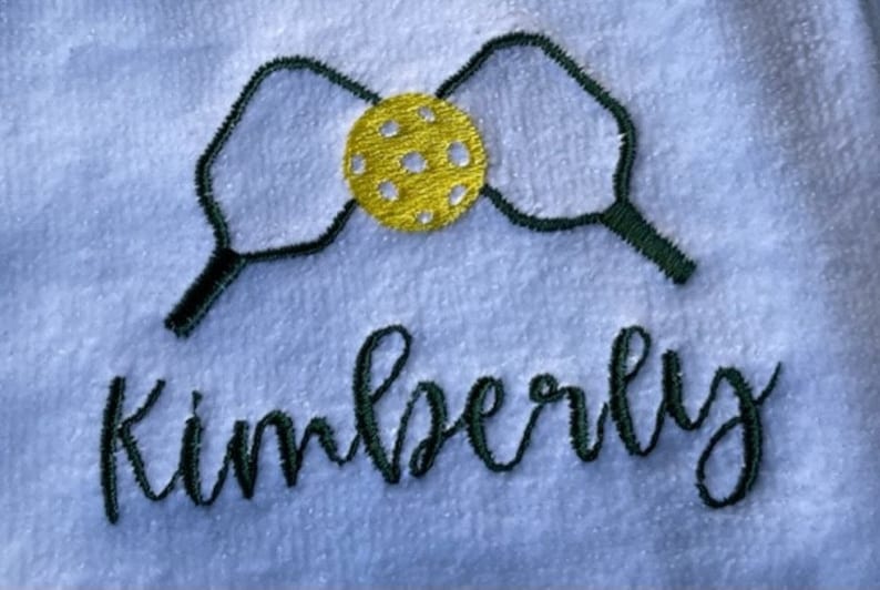 May include: A white towel with green embroidered pickleball paddles and a yellow ball in the center. The name Kimberly is embroidered below the paddles in green.