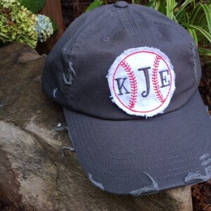 K&ouml;nnte beinhalten: Eine verwaschene, graue Baseballkappe mit einem wei&szlig;-roten, gestickten Baseball-Patch. Der Patch zeigt die Initialen "KJE" in einer Serifenschrift. Die Kappe hat einen gebogenen Schirm und sichtbare Abnutzungserscheinungen.