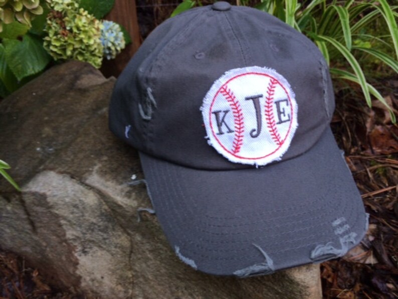 K&ouml;nnte beinhalten: Eine verwaschene, graue Baseballkappe mit einem wei&szlig;-roten, gestickten Baseball-Patch. Der Patch zeigt die Initialen "KJE" in einer Serifenschrift. Die Kappe hat einen gebogenen Schirm und sichtbare Abnutzungserscheinungen.