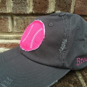 K&ouml;nnte beinhalten: Graue Baseballkappe mit abgenutztem Schirm und einem rosa Baseball-Patch. Das Wort "Brooklyn" ist in Rosa auf die Seite gestickt. Die Kappe hat einen Knopf oben.