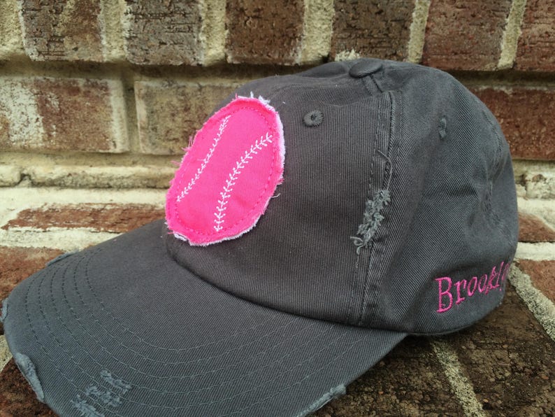 K&ouml;nnte beinhalten: Graue Baseballkappe mit abgenutztem Schirm und einem rosa Baseball-Patch. Das Wort "Brooklyn" ist in Rosa auf die Seite gestickt. Die Kappe hat einen Knopf oben.