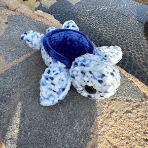 Puede incluir: Un peluche de tortuga hecho a mano en tonos azules y blancos. El caparazón de la tortuga es de un azul intenso y liso, mientras que el cuerpo y las aletas son una mezcla moteada de hilo blanco y azul. El peluche está sobre una superficie de piedra gris.