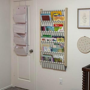 Op de afbeelding: Een aan de muur gemonteerde houten boekenplank met vijf niveaus van grijs en wit gestreepte stoffen vakken met diverse kinderboeken, naast een witte deur met een hangende organizer met vier vakken en ingelijste kunst aan weerszijden.