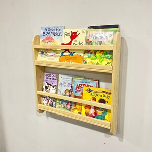 May include: A wooden wall-mounted bookshelf with three shelves, holding various children's books. The books have colorful covers with titles like "A Book for Bramble," "Fox in Socks," and "Alligator Baby." The bookshelf is made of light-colored wood.