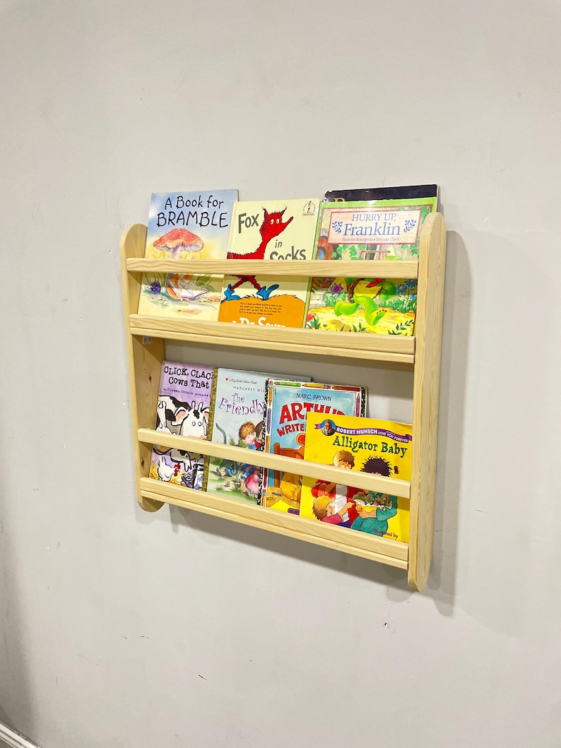 May include: A wooden wall-mounted bookshelf with three shelves, holding various children's books. The books have colorful covers with titles like "A Book for Bramble," "Fox in Socks," and "Alligator Baby." The bookshelf is made of light-colored wood.