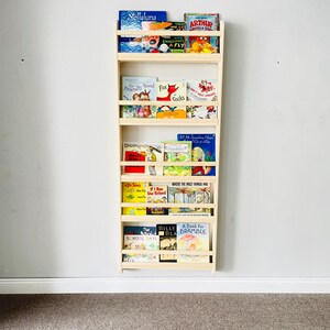 May include: A natural wood wall-mounted bookshelf filled with children's books. The bookshelf has multiple shelves and is mounted on a light gray wall. The books have colorful covers and titles such as "Stellaluna" and "Arthur".