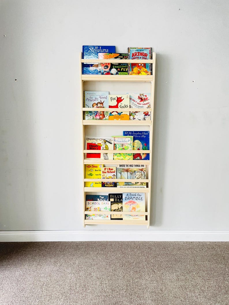 May include: A natural wood wall-mounted bookshelf filled with children's books. The bookshelf has multiple shelves and is mounted on a light gray wall. The books have colorful covers and titles such as "Stellaluna" and "Arthur".