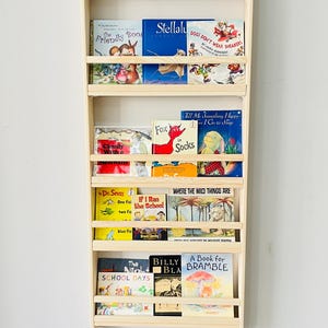May include: A wooden bookshelf filled with colorful children's books. The bookshelf has multiple shelves and is mounted on a light gray wall. Titles include "Hurry Up Franklin", "Arthur Writes a Story", and "Where the Wild Things Are".