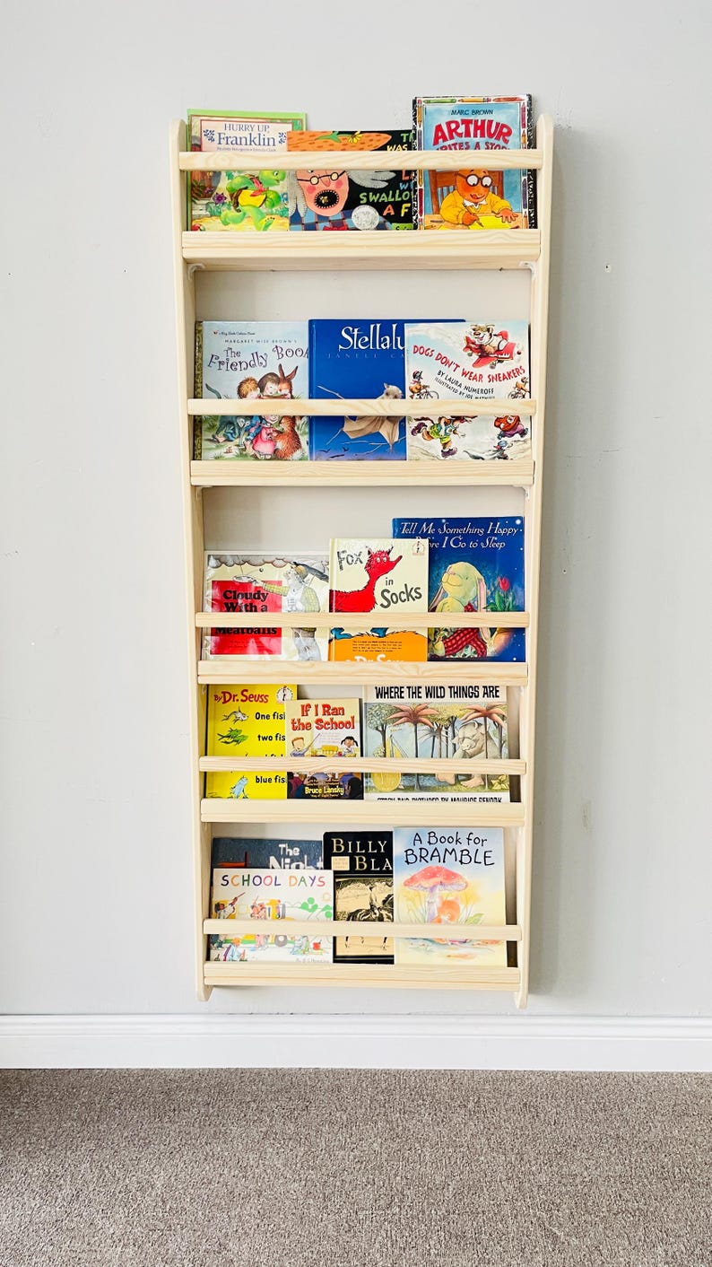 May include: A wooden bookshelf filled with colorful children's books. The bookshelf has multiple shelves and is mounted on a light gray wall. Titles include "Hurry Up Franklin", "Arthur Writes a Story", and "Where the Wild Things Are".