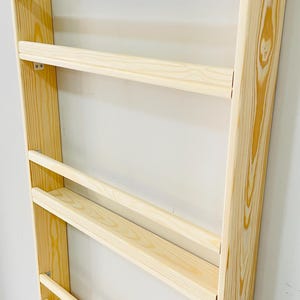 May include: A wooden wall-mounted bookshelf with a light natural finish. The bookshelf has a ladder-like design with multiple horizontal shelves. The wood grain is visible, and the shelves are evenly spaced.
