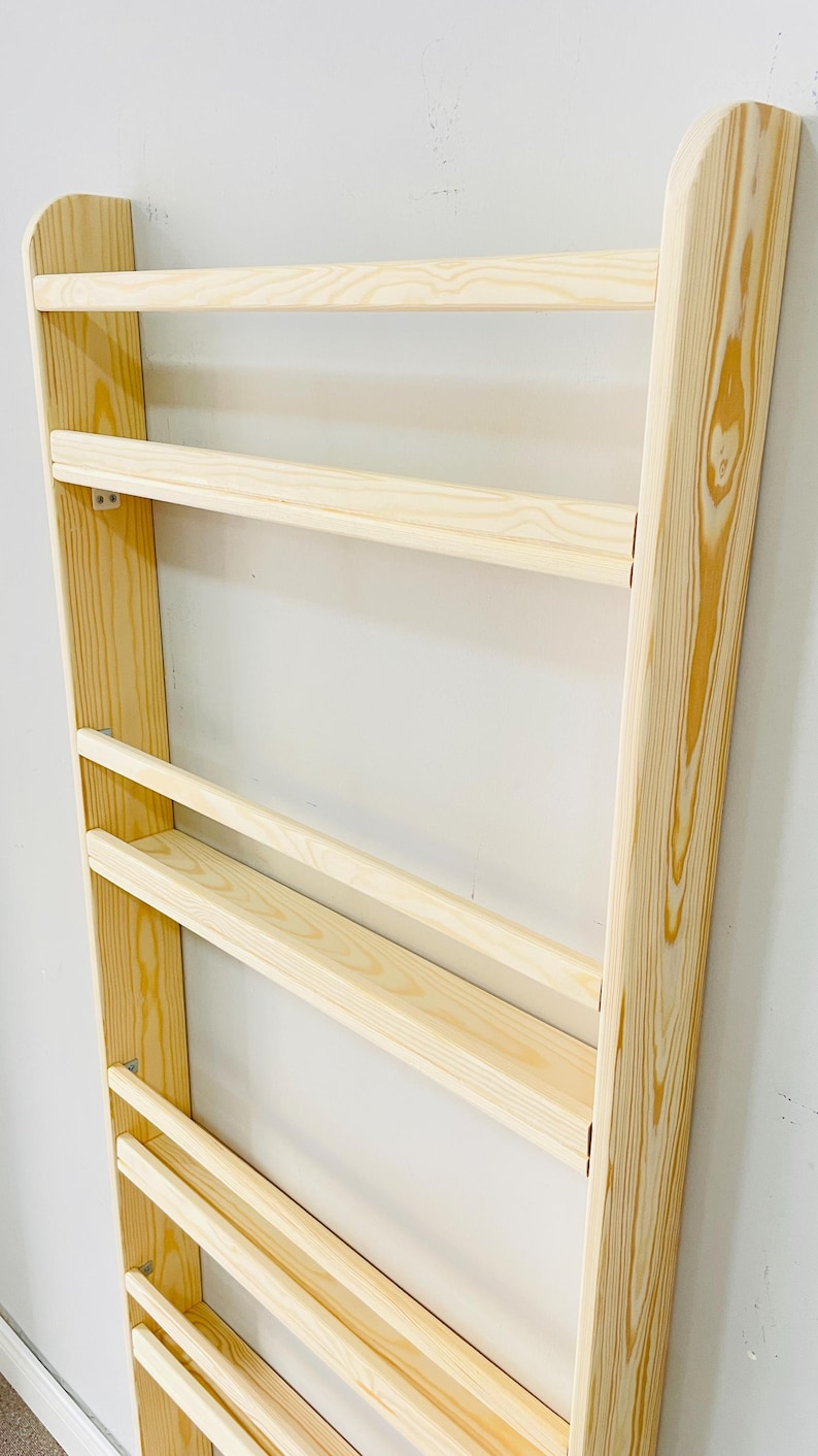May include: A wooden wall-mounted bookshelf with a light natural finish. The bookshelf has a ladder-like design with multiple horizontal shelves. The wood grain is visible, and the shelves are evenly spaced.