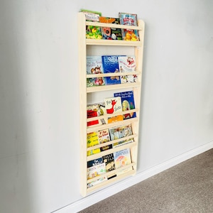 May include: A light-colored wooden bookshelf mounted on a wall, filled with children's books. The bookshelf has multiple shelves to display the books. The books have colorful covers and various titles. The bookshelf is made of natural wood.