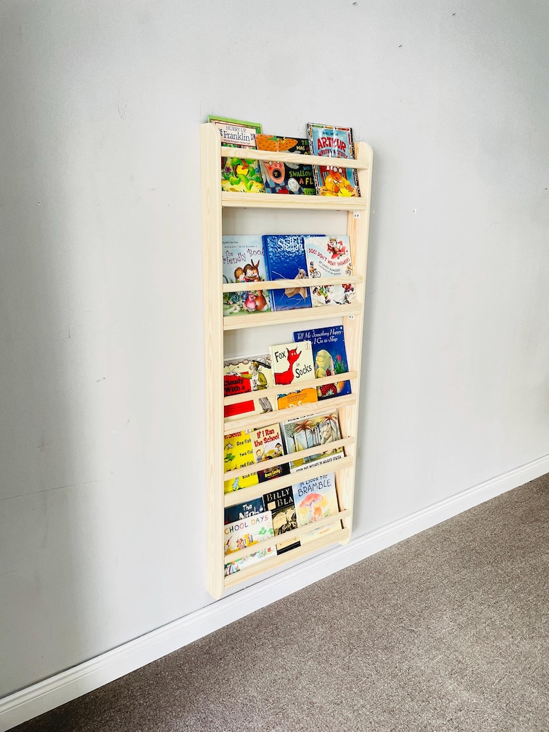 May include: A light-colored wooden bookshelf mounted on a wall, filled with children's books. The bookshelf has multiple shelves to display the books. The books have colorful covers and various titles. The bookshelf is made of natural wood.