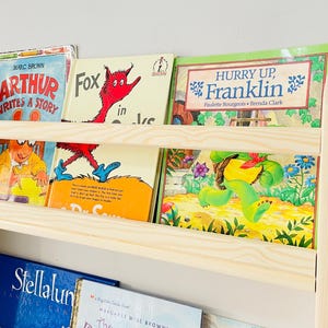 May include: A light-colored wooden bookshelf filled with children's books. The books have colorful covers with titles like "Arthur Writes a Story," "Fox in Socks," and "Hurry Up, Franklin." The bookshelf has two shelves and is mounted on a white wall.