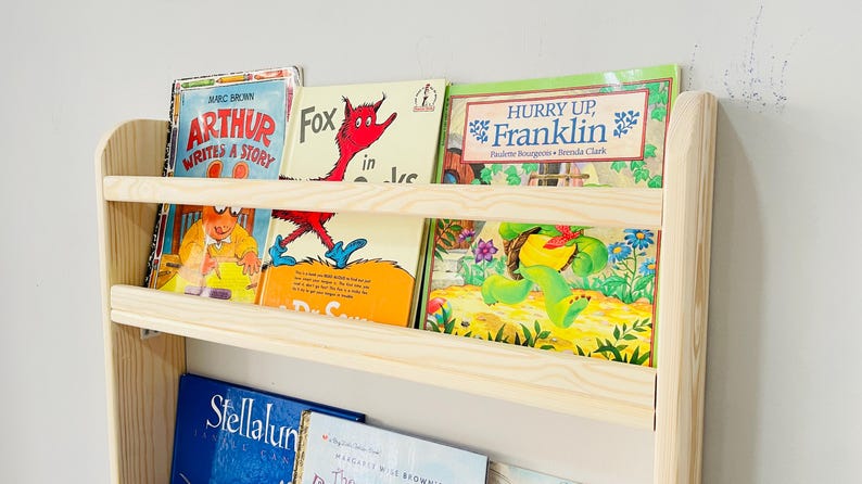 May include: A light-colored wooden bookshelf filled with children's books. The books have colorful covers with titles like "Arthur Writes a Story," "Fox in Socks," and "Hurry Up, Franklin." The bookshelf has two shelves and is mounted on a white wall.