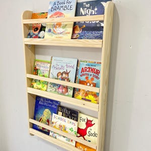May include: A wooden wall-mounted bookshelf filled with children's books. The bookshelf is made of light-colored wood and has three shelves. The books have colorful covers and various titles, including "A Book for Bramble" and "Arthur Writes a Story".
