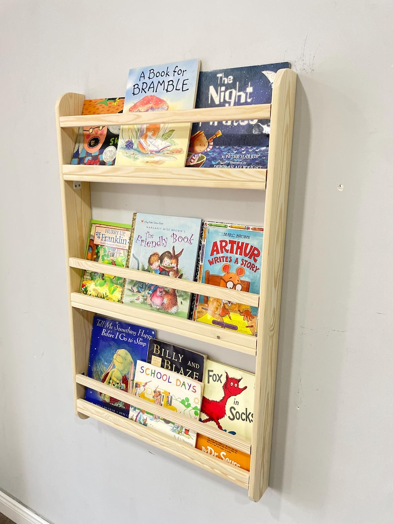 May include: A wooden wall-mounted bookshelf filled with children's books. The bookshelf is made of light-colored wood and has three shelves. The books have colorful covers and various titles, including "A Book for Bramble" and "Arthur Writes a Story".
