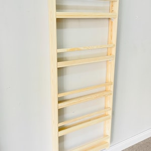 May include: A light-colored wooden wall-mounted bookshelf with multiple horizontal shelves. The bookshelf is made of unfinished wood and is attached to a light gray wall. The shelves are evenly spaced and designed for storage.