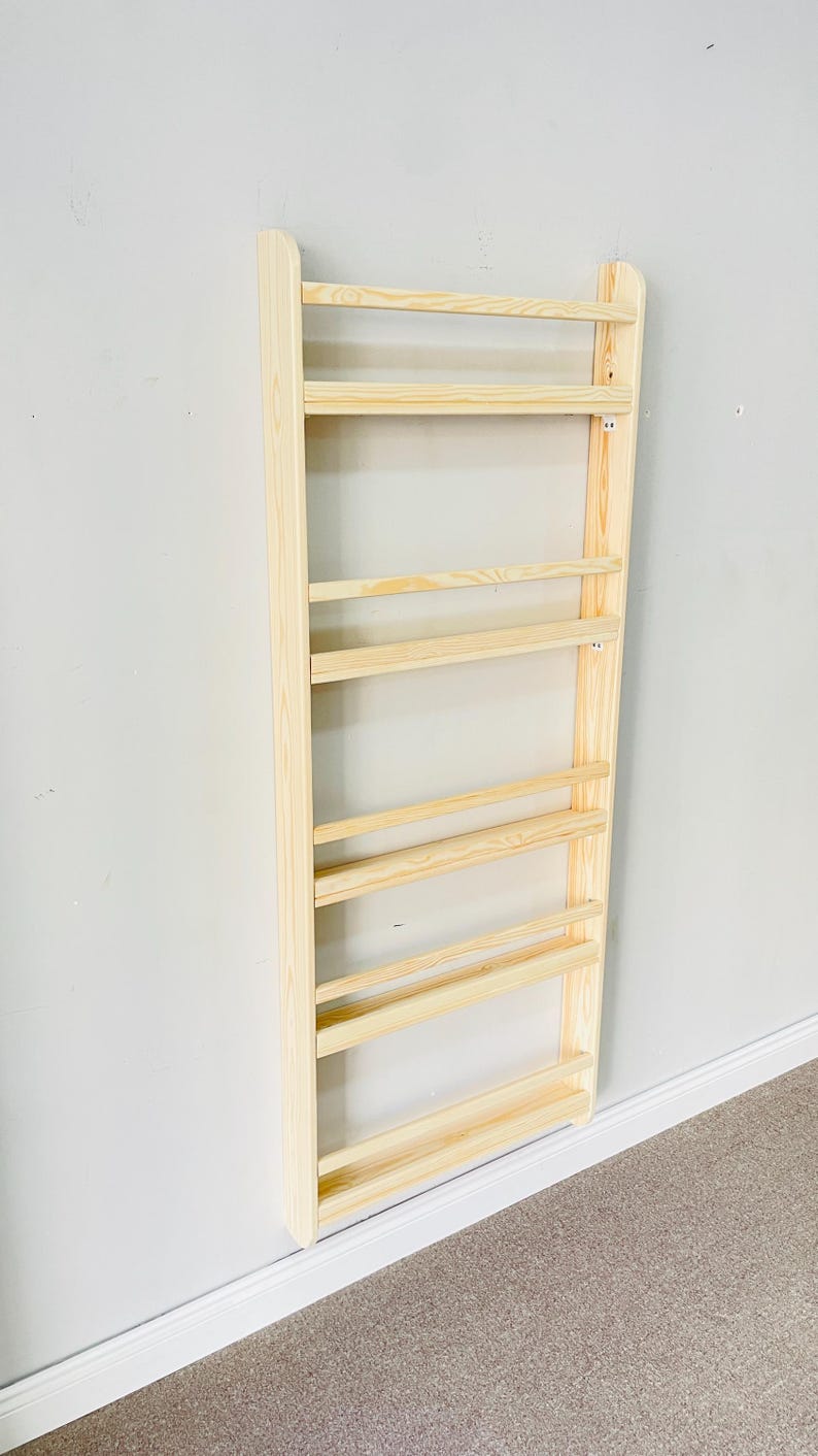 May include: A light-colored wooden wall-mounted bookshelf with multiple horizontal shelves. The bookshelf is made of unfinished wood and is attached to a light gray wall. The shelves are evenly spaced and designed for storage.