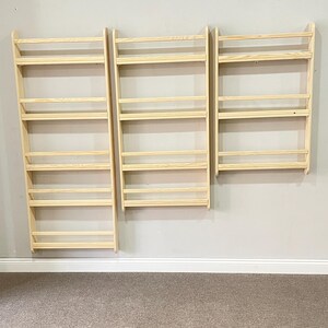 May include: Three unfinished wooden wall-mounted spice racks. Each rack has multiple horizontal shelves and a simple, minimalist design. The racks are mounted on a light gray wall above a beige carpeted floor.