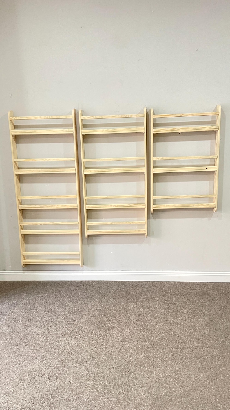 May include: Three unfinished wooden wall-mounted spice racks. Each rack has multiple horizontal shelves and a simple, minimalist design. The racks are mounted on a light gray wall above a beige carpeted floor.