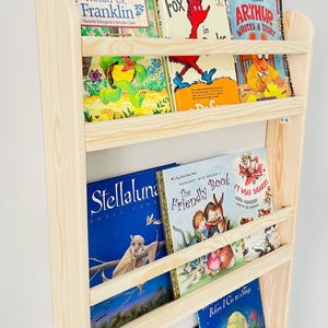 May include: A wooden bookshelf filled with colorful children's books. Titles include "Hurry Up, Franklin", "Fox in Socks", "Arthur Writes a Story", and "Stellaluna". The bookshelf is made of light-colored wood and is mounted on a white wall.